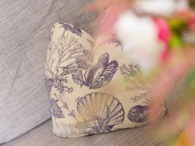 A pillow with shell and seaweed patterns on a grey fabric sofa with blurred flowers in the foreground at Daphne in Mullion