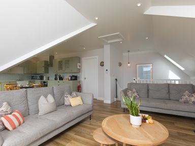 A living room with two gray sofas a round wooden coffee table with a plant and snacks an open kitchen with green cabinets and a dining table at Daphne in Mullion