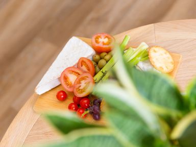 A wooden board with slices of brie cheese tomatoes green olives celery crackers and grapes on a wooden table