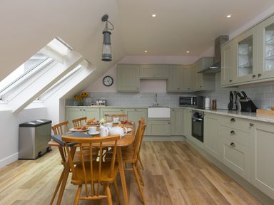 A kitchen with wooden dining table and chairs set with breakfast dishes under sloped windows at Daphne in Mullion