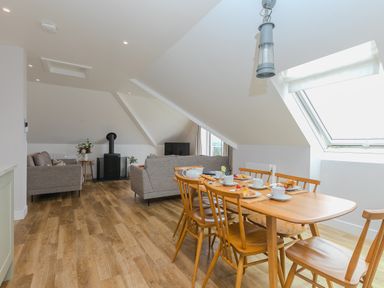 A dining area with wooden table and chairs set with teacups and pastries next to a living area with grey sofas and a wood burner at Daphne in Mullion