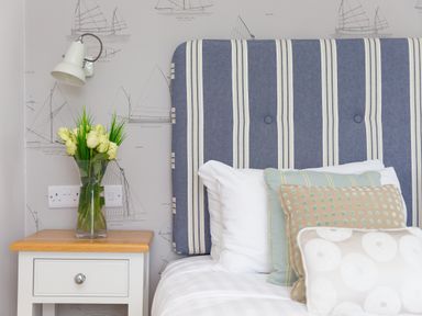 A bedroom with a striped headboard pillows a bedside table with a vase of white roses and a wall lamp at Daphne in Mullion