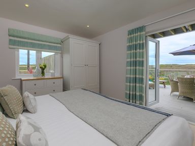 A bedroom with a bed pillows and blanket a white dresser with flowers a wardrobe and a door opening to a patio with outdoor furniture at Daphne in Mullion