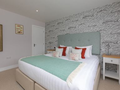 A bedroom with a bed with a teal throw and red and white pillows two nightstands with lamps and fish patterned wallpaper at Daphne in Mullion