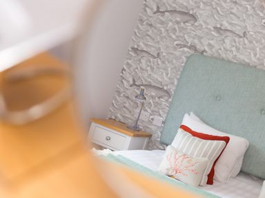 A bed with pillows and a blue headboard next to a white nightstand with a lamp and wallpaper with fish in a bedroom at Daphne in Mullion