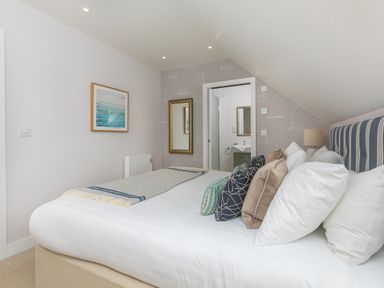 A bedroom with a bed, pillows, a blanket, wall art, a mirror, and a door leading to a bathroom at Daphne in Mullion