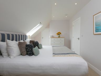 A bedroom with a bed with multiple pillows and a blanket a chest of drawers with a vase of sunflowers and a painting on the wall at Daphne in Mullion