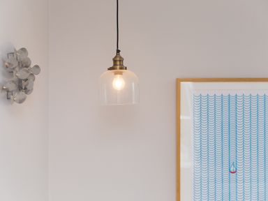 A wall with a hanging light bulb, a metal wall decoration, and a framed print of a blue wave pattern with a small red sailboat at Daphne in Mullion