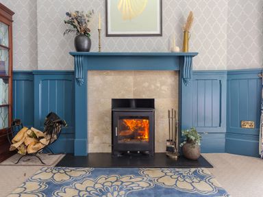 A fireplace with a burning wood stove and blue painted wood paneling at Daphne in Mullion