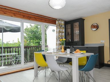 A dining room with a white table and colorful chairs near glass sliding doors and a black cabinet at 37 Tamar in St Ann's Chapel near Callington