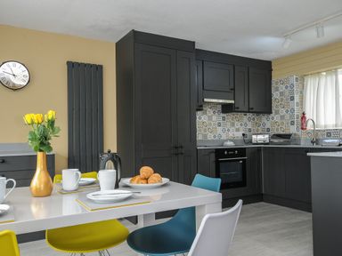 A kitchen with dark cabinets a tiled backsplash a dining table with plates mugs croissants and a vase of yellow flowers at 37 Tamar in St Ann's Chapel near Callington
