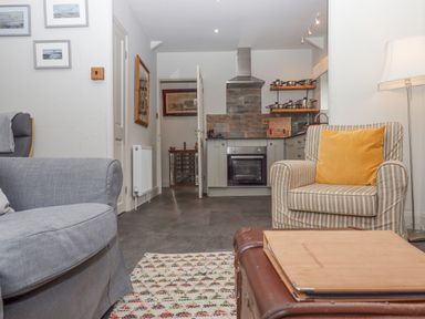 A living room with a grey sofa a striped armchair with a yellow cushion a lamp and kitchen area with oven and pots at Hole Farm Stables in Bickington near Ashburton
