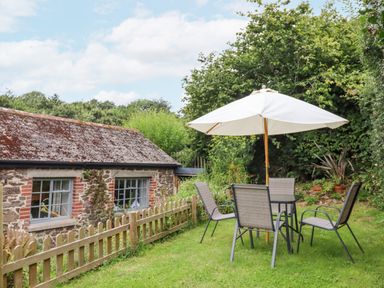 A garden with a stone building with windows a wooden fence and an outdoor table with four chairs and a white umbrella at Hole Farm Stables in Bickington near Ashburton