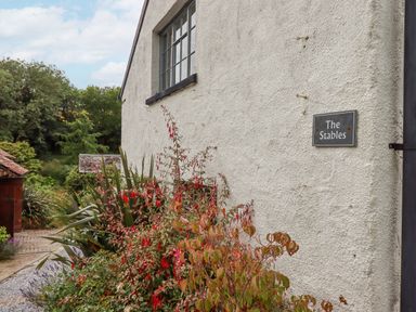 Exterior wall of a white building with a window and a sign that says The Stables with plants and flowers alongside a pathway at Hole Farm Stables Bickington near Ashburton