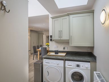 A utility room with washing machine and dryer under cabinets and a dining area visible beyond at Leylands in Salcombe