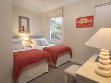 A bedroom with two single beds a bedside table with a lamp a wall mirror a window a desk with a lamp and a stool at Leylands in Salcombe