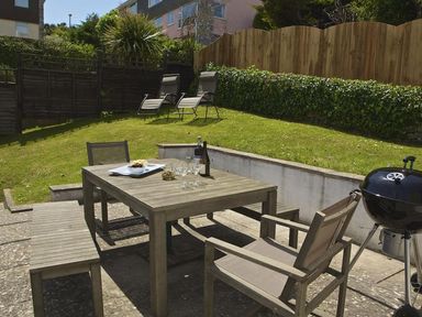 An outdoor patio with a wooden table and chairs a barbecue and two lounge chairs on a grassy area at Leylands in Salcombe