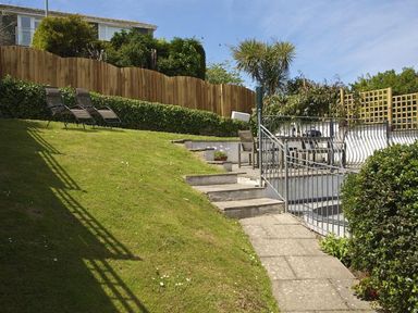 A garden with a grassy slope two lounge chairs a stone pathway stairs metal railing and outdoor seating area at Leylands in Salcombe