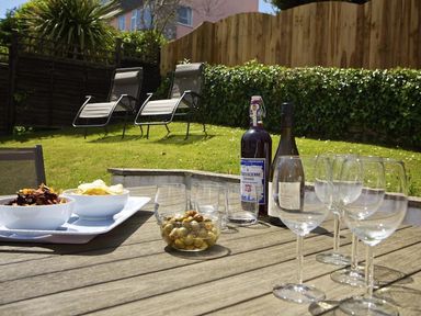 An outdoor wooden table with wine glasses olives bottles and bowls of snacks in a garden with lounge chairs at Leylands in Salcombe