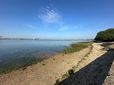 A shoreline with a pebble beach and calm water with trees and industrial structures in the distance at Inishnee House in Shotley Gate
