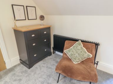 A corner of a room with a dark chest of drawers topped with framed art and a mirror and a brown chair with a patterned cushion near a black radiator at The Grove in Wolfsdale near Haverfordwest