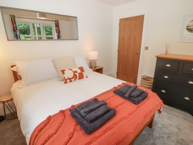 A bedroom with a bed covered by white sheets and an orange blanket with towels on top a bedside table with a lamp a black dresser a wooden door and a mirror reflecting a window at The Grove in Wolfsdale near Haverfordwest