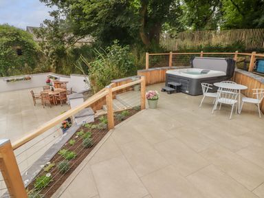 An outdoor patio area with a hot tub and white metal chairs and table beside a wooden railing with plants and a lower patio with wooden chairs and tables surrounded by trees at The Grove in Wolfsdale near Haverfordwest