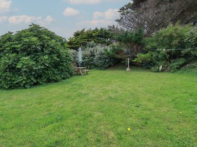 A grassy garden with bushes trees a picnic table with an umbrella and a bird feeder at Horseshoe Cottage Near Holywell Bay