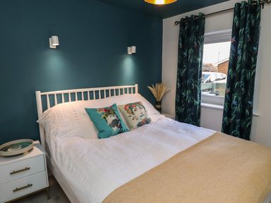 A bedroom with a white bed and two decorated pillows against a blue wall with two wall lights and a window with patterned curtains at Villa Rose in Beadnell near Seahouses