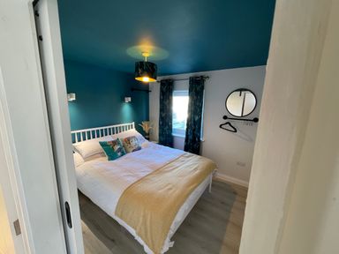 A bedroom with a double bed white frame pillows a yellow blanket patterned curtains and a round mirror on the wall at Villa Rose in Beadnell near Seahouses