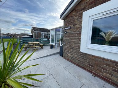 A patio area with a wooden picnic table and benches a potted plant and a brick house with a window at Villa Rose Beadnell near Seahouses