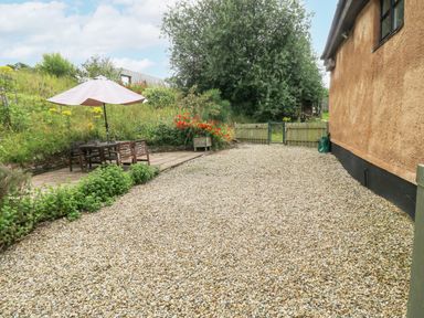 An outdoor patio area with a gravel ground a wooden table with chairs and an umbrella at Ixworth Cottage in Little Woodford near Ottery St Mary