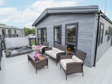 An outdoor patio with wicker furniture a glass table a hot tub and flowers at Sunset Lodge in Woodhall Spa