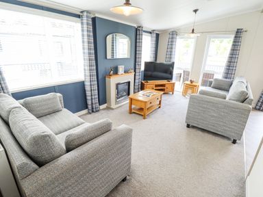 A living room with two grey sofas a wooden coffee table a television on a wooden stand a fireplace and windows with plaid curtains at Sunset Lodge in Woodhall Spa