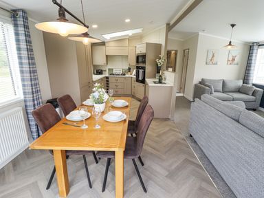 A dining table with chairs set for four near a kitchen and a living area with sofas and wall art at Sunset Lodge in Woodhall Spa
