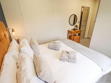 A bedroom with a bed with white and beige pillows and folded towels on top with a wooden dressing table with a round mirror and a door in the background at Sunset Lodge in Woodhall Spa