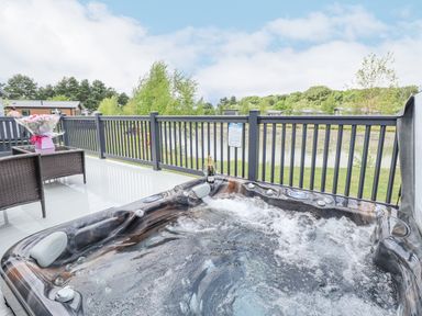 A bubbling hot tub on a deck with a bottle of champagne and two glasses and two wicker chairs with a table holding flowers at Sunset Lodge in Woodhall Spa