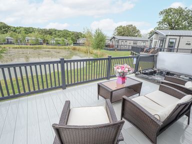 A deck with wicker chairs a table with flowers and a hot tub overlooking a pond and holiday homes at Sunset Lodge in Woodhall Spa