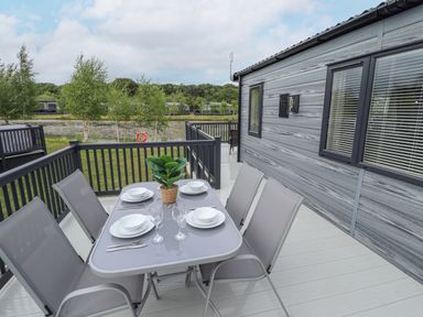 An outdoor dining table set with plates glasses and cutlery on a deck beside a grey wooden house with trees and a pond in the background at Sunset Lodge in Woodhall Spa