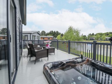 An outdoor deck with wicker chairs a table a hot tub and a view of a pond at Sunset Lodge in Woodhall Spa