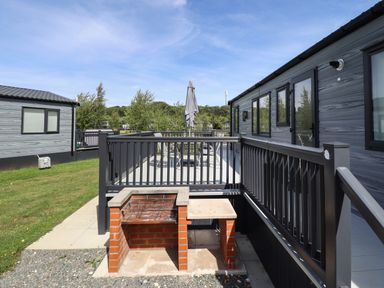 An outdoor patio with a brick barbecue grill and a seating area beside grey modern lodges at Sunset Lodge in Woodhall Spa