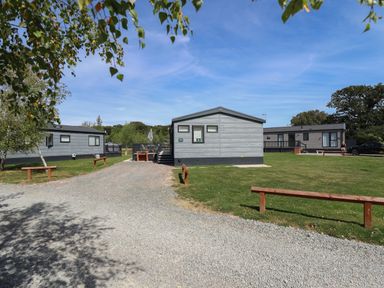 Three gray cabins on grass with gravel pathway and wooden benches at Sunset Lodge in Woodhall Spa