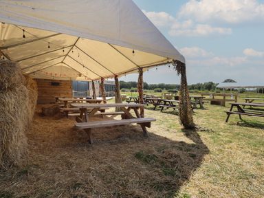 An outdoor picnic area with wooden benches under a tent and hay bales surrounding the space at Sunset Lodge in Woodhall Spa