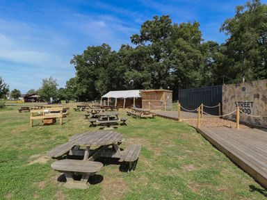 An outdoor seating area with wooden picnic tables and a wooden food stall under trees at Sunset Lodge in Woodhall Spa