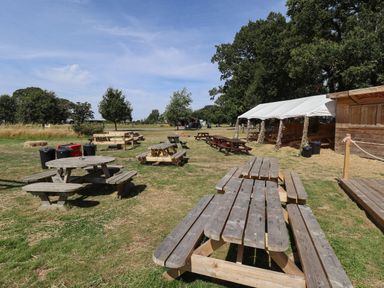 An outdoor seating area with wooden picnic tables and benches near a tent and wooden shelter in a grassy field