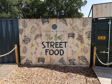 A wooden sign with illustrations of food and text saying country kitchen the best street food outside surrounded by trees