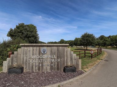 A wooden sign reading Woodhall Country Park beside a road with trees and a speed limit sign in Woodhall Spa