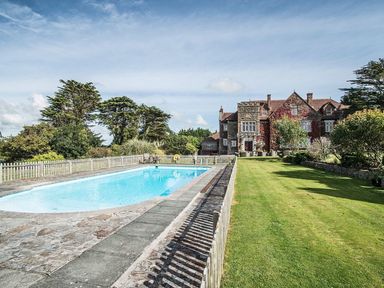An outdoor swimming pool with a stone patio and a large stone house with greenery in the background at Alston Hall in Battisborough Cross