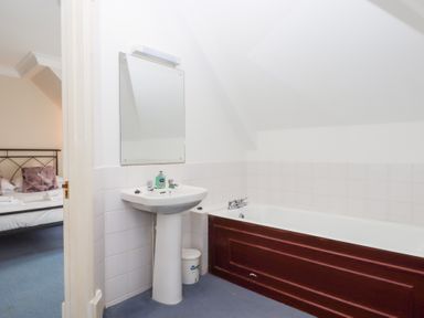 A bathroom with a sink, mirror, bathtub with wooden panel and a glimpse of a bedroom at Alston Hall in Battisborough Cross