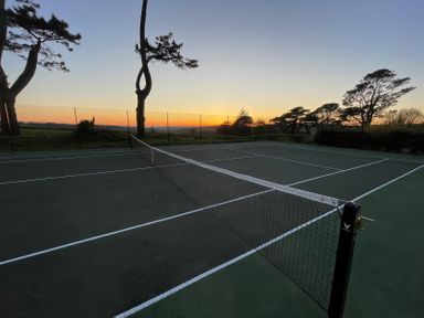 An empty outdoor tennis court with a net at sunset surrounded by trees and a wire fence at Alston Hall in Battisborough Cross